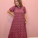 A woman modeling a short-sleeve button-up maxi dress with a pink and brown block print pattern, standing against a solid pink wall.