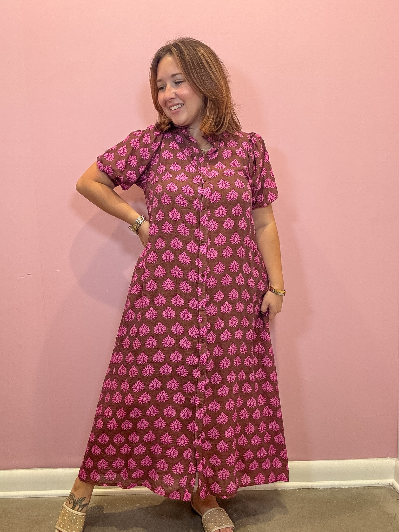 A woman modeling a short-sleeve button-up maxi dress with a pink and brown block print pattern, standing against a solid pink wall.