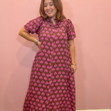 A woman modeling a short-sleeved, button-up maxi dress with a pink and brown block print pattern, standing against a solid pink wall.