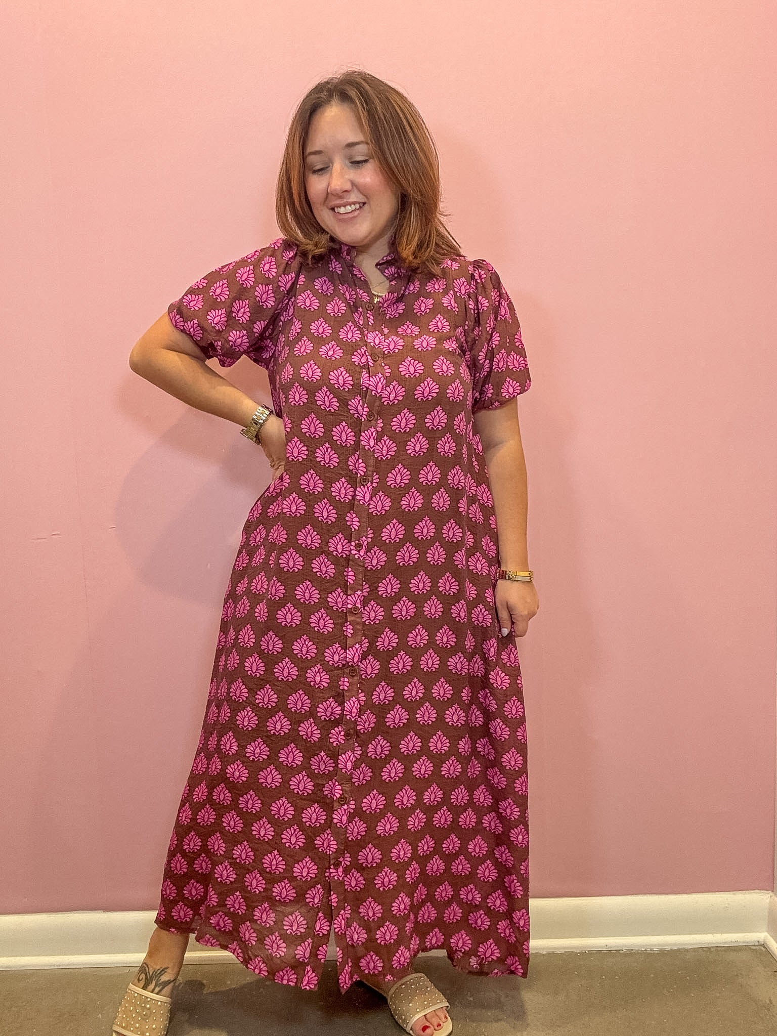 A woman modeling a short-sleeved, button-up maxi dress with a pink and brown block print pattern, standing against a solid pink wall.