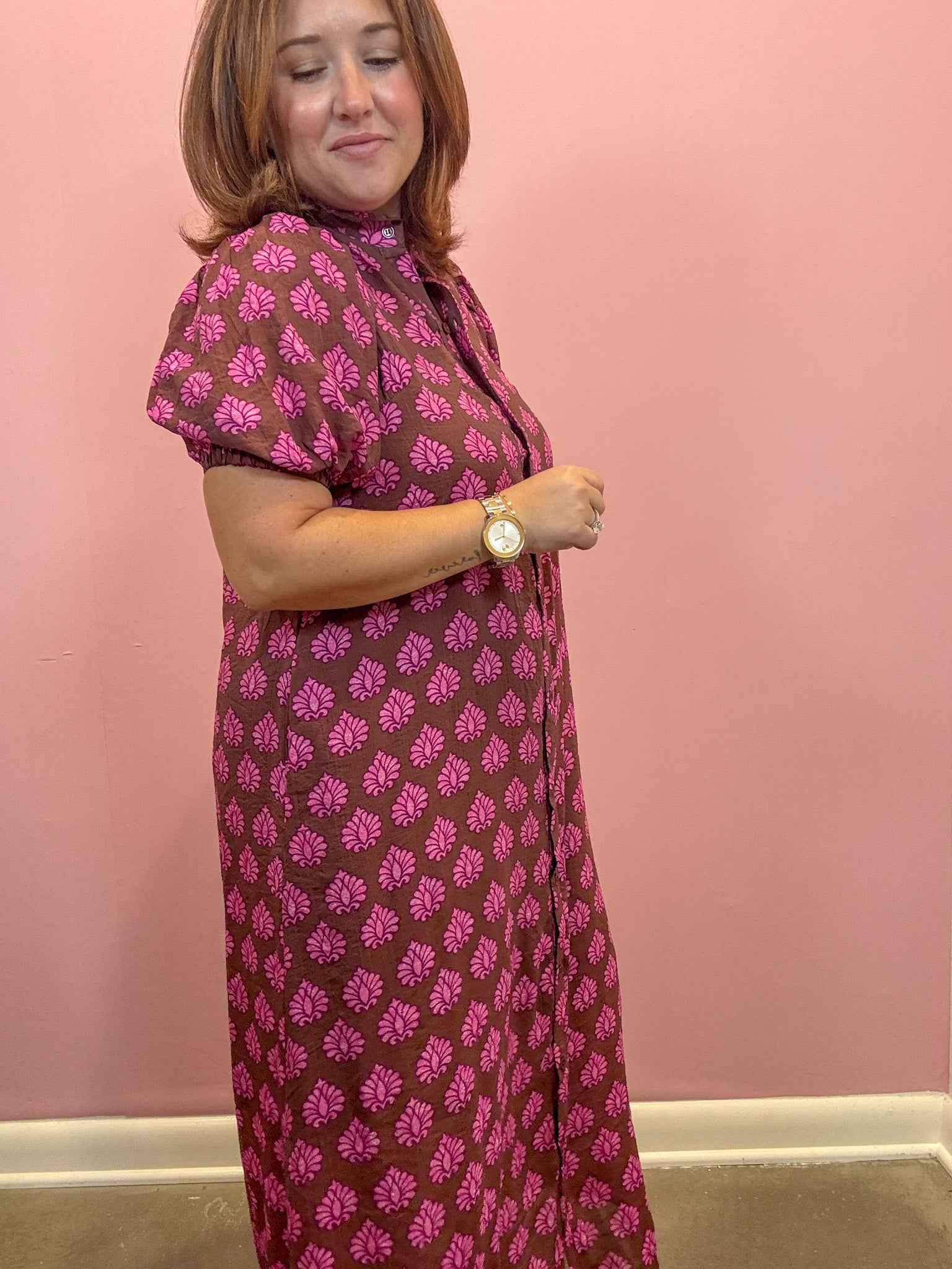 A woman modeling a brown and pink patterned dress against a solid pink wall.
