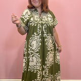 A woman wearing a short-sleeved, knee-length dress with an olive green and cream damask pattern, standing against a solid pink wall.