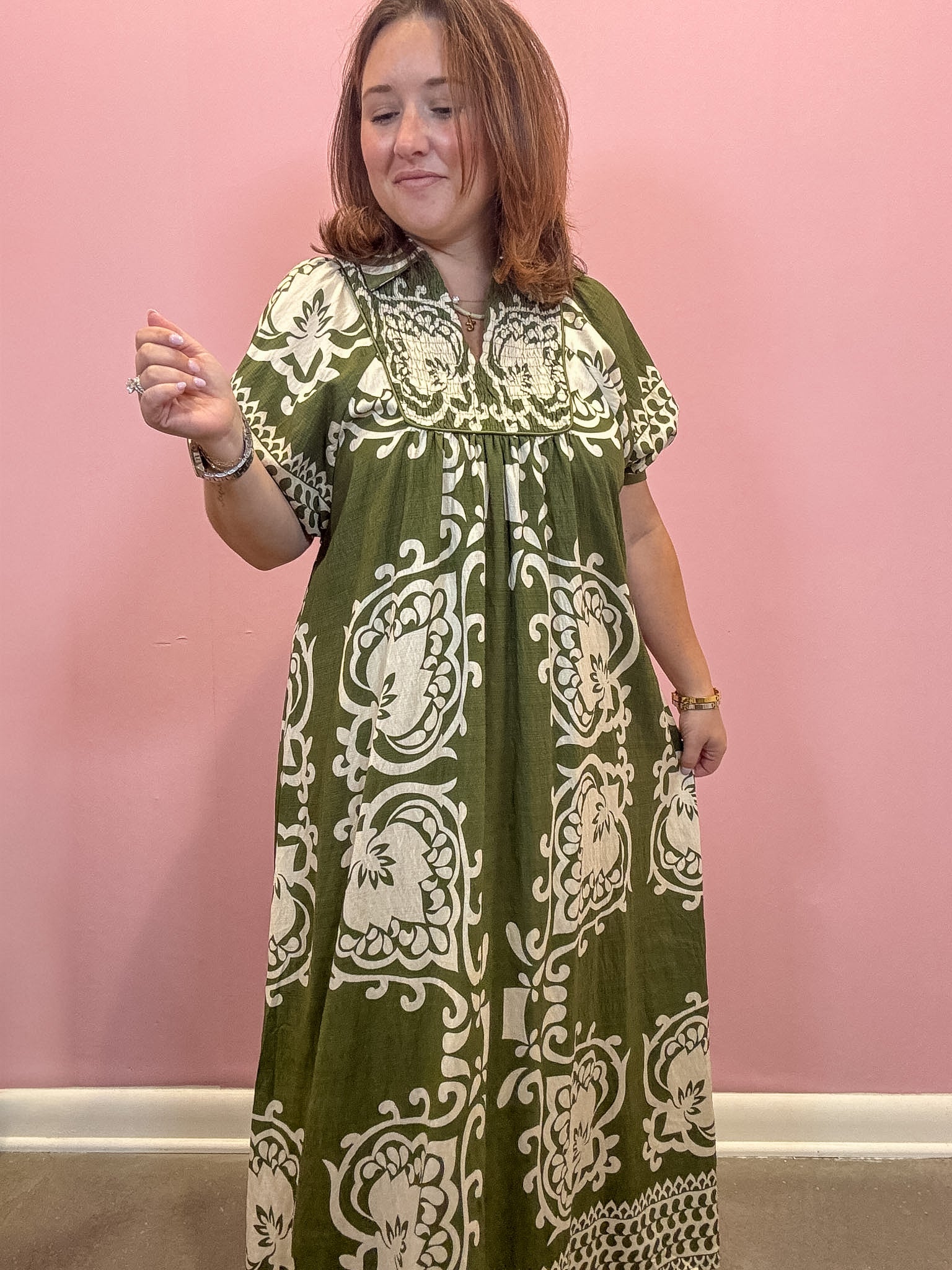 A woman wearing a short-sleeved, knee-length dress with an olive green and cream damask pattern, standing against a solid pink wall.