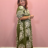 A woman modeling a green and cream patterned maxi dress against a solid pink wall.