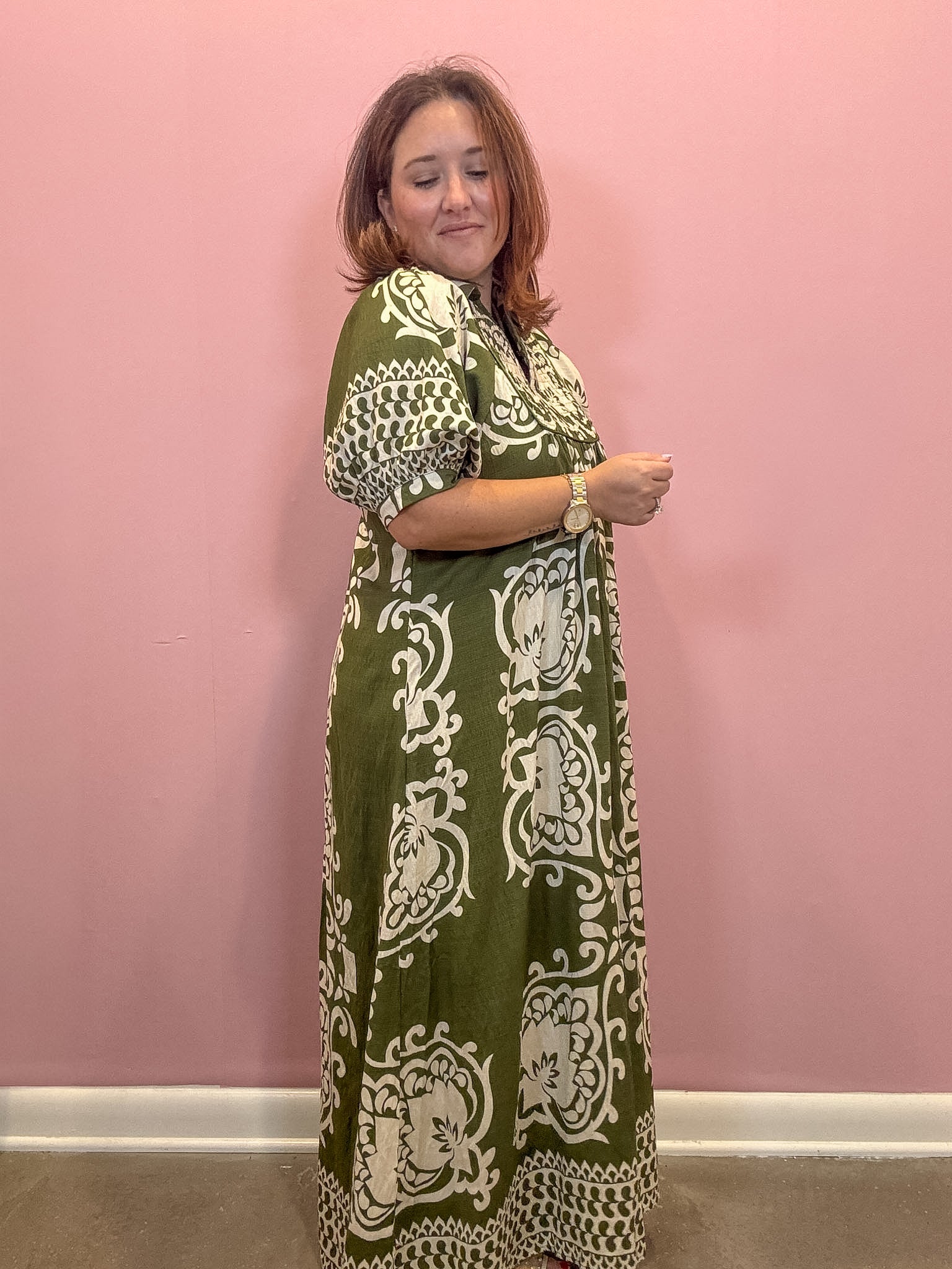 A woman modeling a green and cream patterned maxi dress against a solid pink wall.