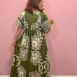 A woman wearing a green and white patterned maxi dress, shown from the back against a solid pink wall.