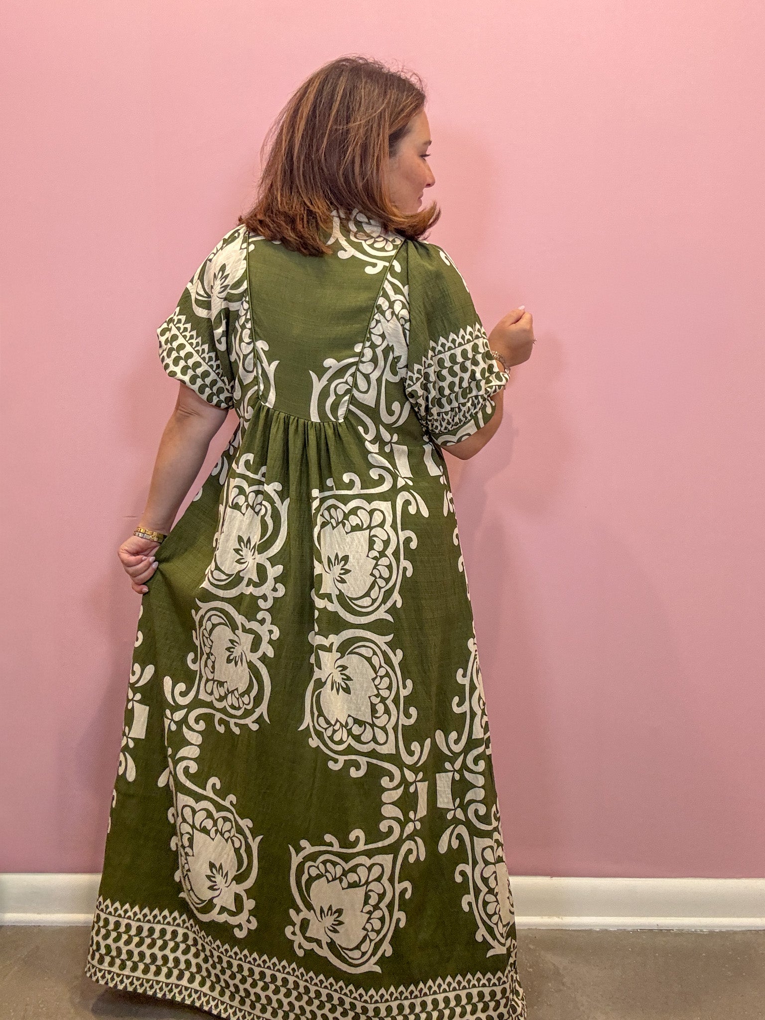 A woman wearing a green and white patterned maxi dress, shown from the back against a solid pink wall.