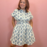 A woman modeling a white short-sleeve button-up dress with a blue floral block print, standing against a solid pink wall.