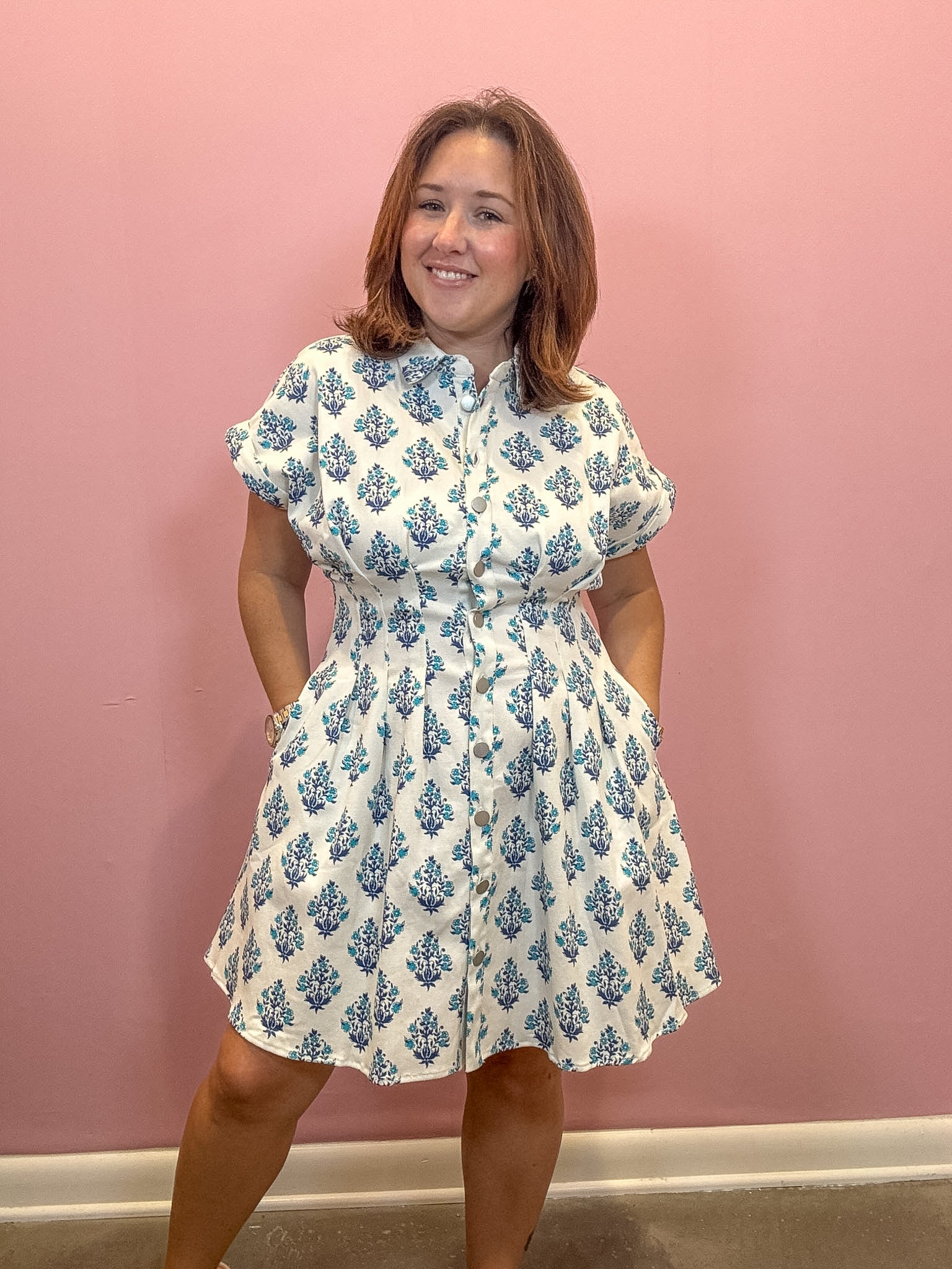 A woman modeling a white short-sleeve button-up dress with a blue floral block print, standing against a solid pink wall.