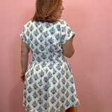 A woman wearing a white short-sleeve dress with a blue floral block print pattern, shown from the back against a solid pink wall.