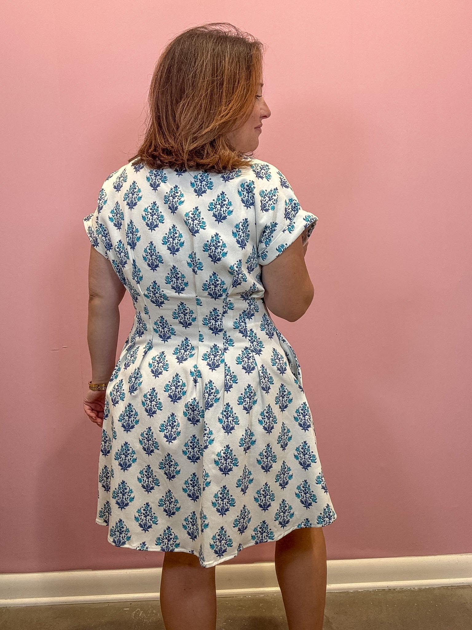 A woman wearing a white short-sleeve dress with a blue floral block print pattern, shown from the back against a solid pink wall.