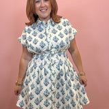 A woman wearing a white short-sleeve button-up dress with a blue floral block print pattern, standing against a solid pink wall.