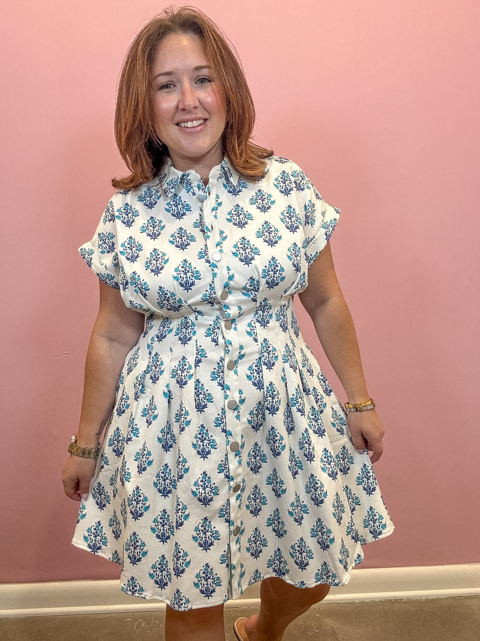 A woman wearing a white short-sleeve button-up dress with a blue floral block print pattern, standing against a solid pink wall.