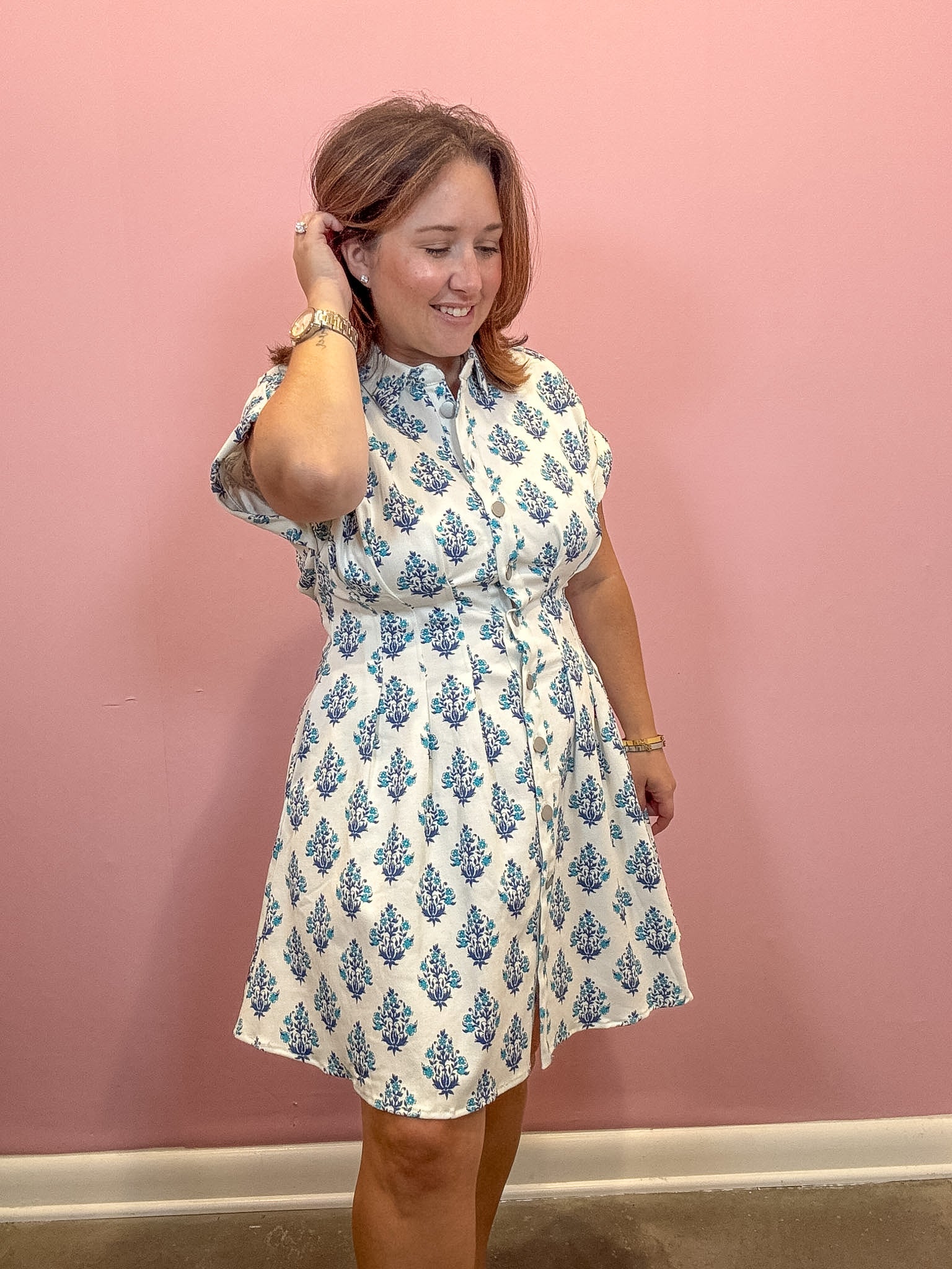 A woman modeling a short-sleeve white button-up dress with a blue floral block print pattern, standing against a solid pink wall.