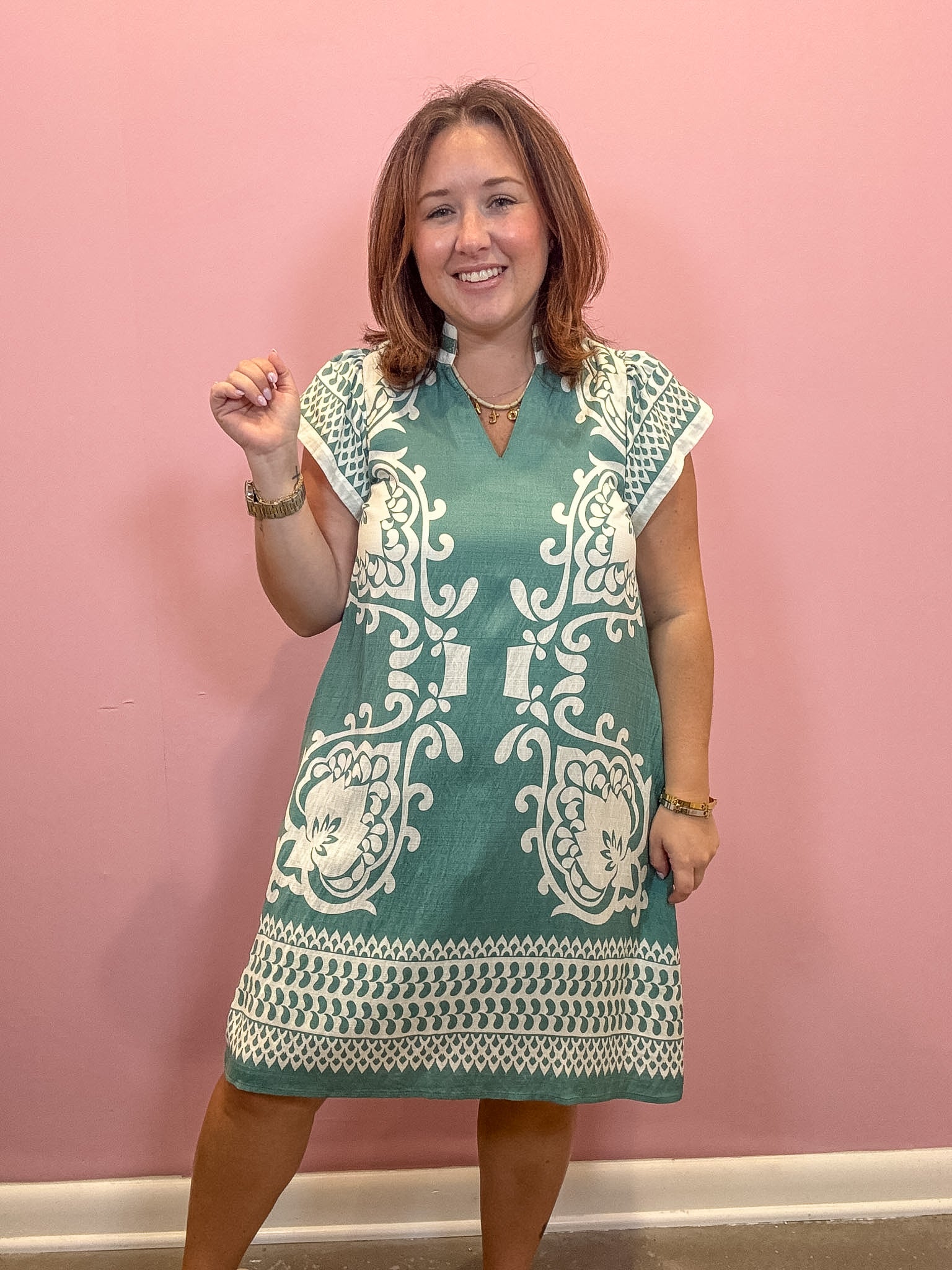 A woman modeling a teal and white patterned short-sleeve tunic dress against a solid pink wall.