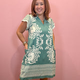 A woman modeling a teal and white patterned short-sleeve tunic dress against a solid pink wall.