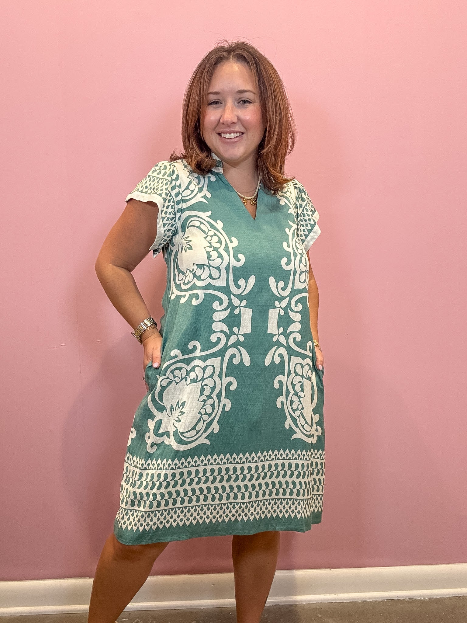 A woman modeling a teal and white patterned short-sleeve tunic dress against a solid pink wall.
