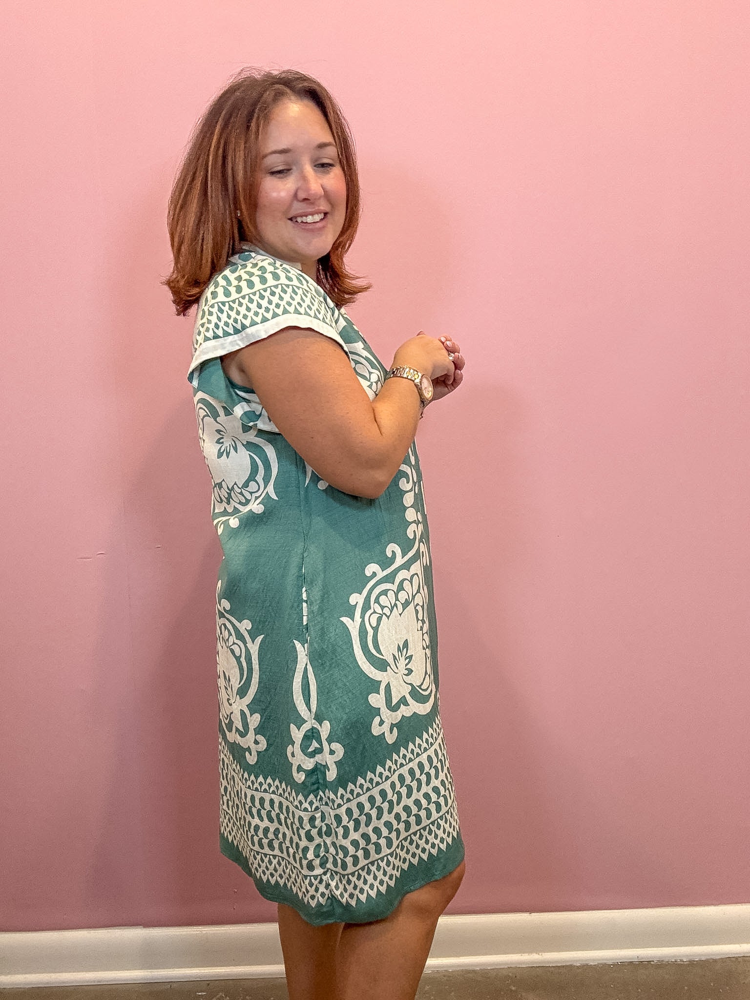 A woman wearing a teal and white patterned sleeveless dress, standing against a solid pink wall.