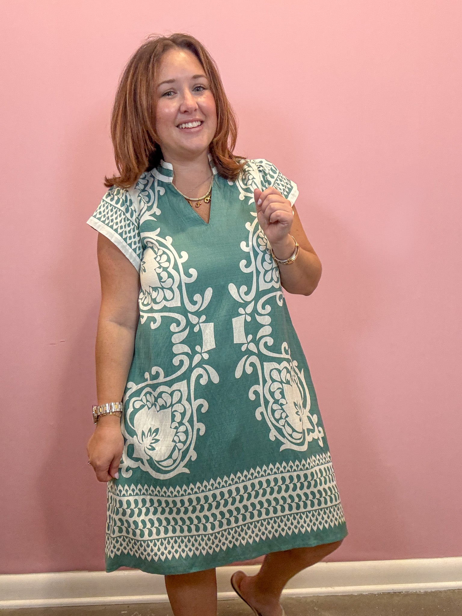 A woman modeling a teal green and white printed tunic dress against a solid pink wall.