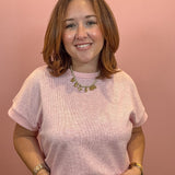 A woman with shoulder-length brown hair smiling, wearing a light pink short-sleeve knit top and sage green pants against a solid dusty pink background.