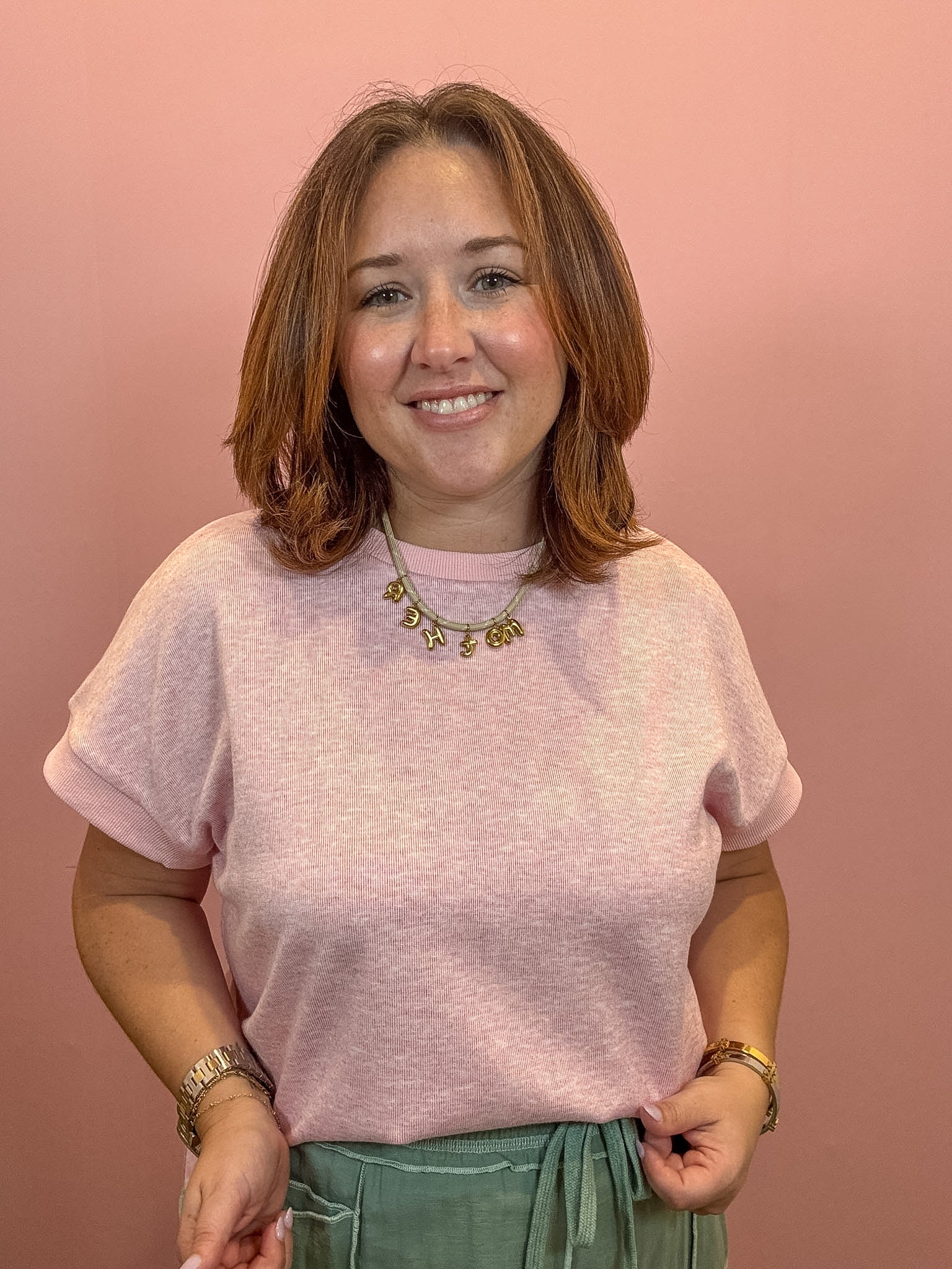 A woman with shoulder-length brown hair smiling, wearing a light pink short-sleeve knit top and sage green pants against a solid dusty pink background.
