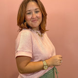 A woman with shoulder-length auburn hair is shown from the waist up, wearing a light pink short-sleeve knit top and sage green drawstring pants, standing against a solid dusty pink wall.