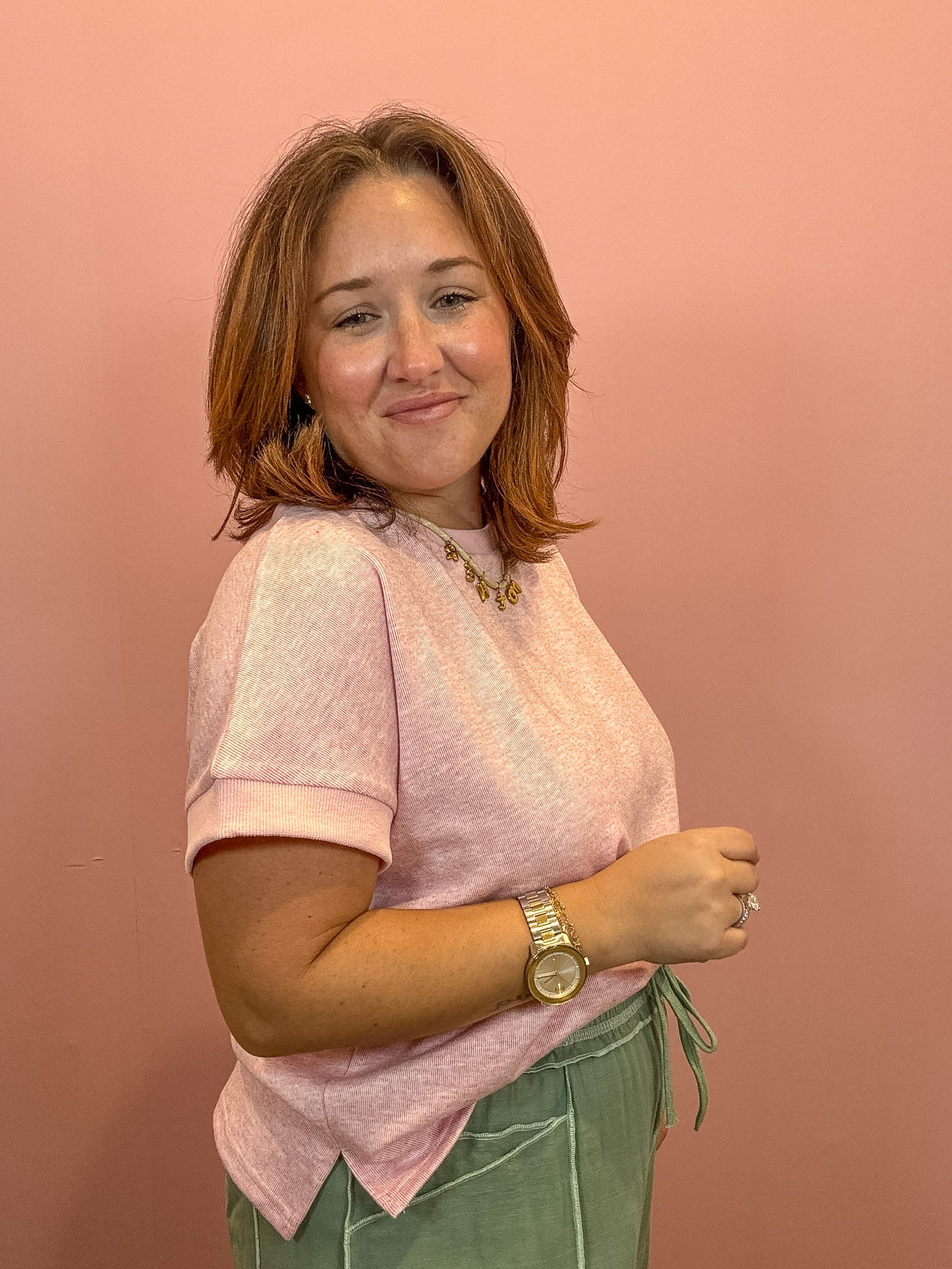 A woman with shoulder-length auburn hair is shown from the waist up, wearing a light pink short-sleeve knit top and sage green drawstring pants, standing against a solid dusty pink wall.