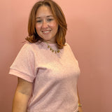 A woman with shoulder-length brown hair smiling, wearing a light pink short-sleeve crewneck t-shirt and olive green pants, standing against a solid dusty pink wall.
