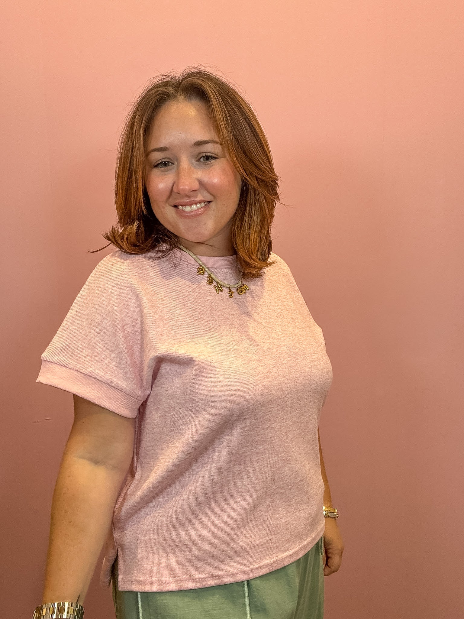A woman with shoulder-length brown hair smiling, wearing a light pink short-sleeve crewneck t-shirt and olive green pants, standing against a solid dusty pink wall.