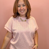 A smiling woman with shoulder-length brown hair wearing a light pink short-sleeve crewneck t-shirt and sage green pants, standing against a solid dusty pink background.