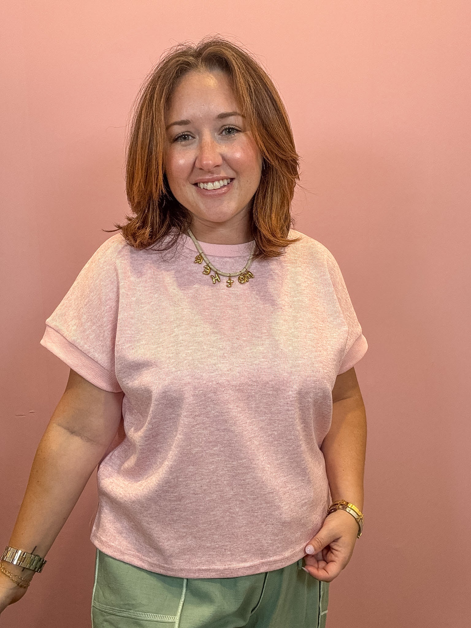 A smiling woman with shoulder-length brown hair wearing a light pink short-sleeve crewneck t-shirt and sage green pants, standing against a solid dusty pink background.