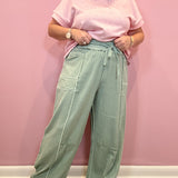 A person wearing a light pink short-sleeve t-shirt and sage green wide-leg drawstring pants, standing against a solid pink wall.