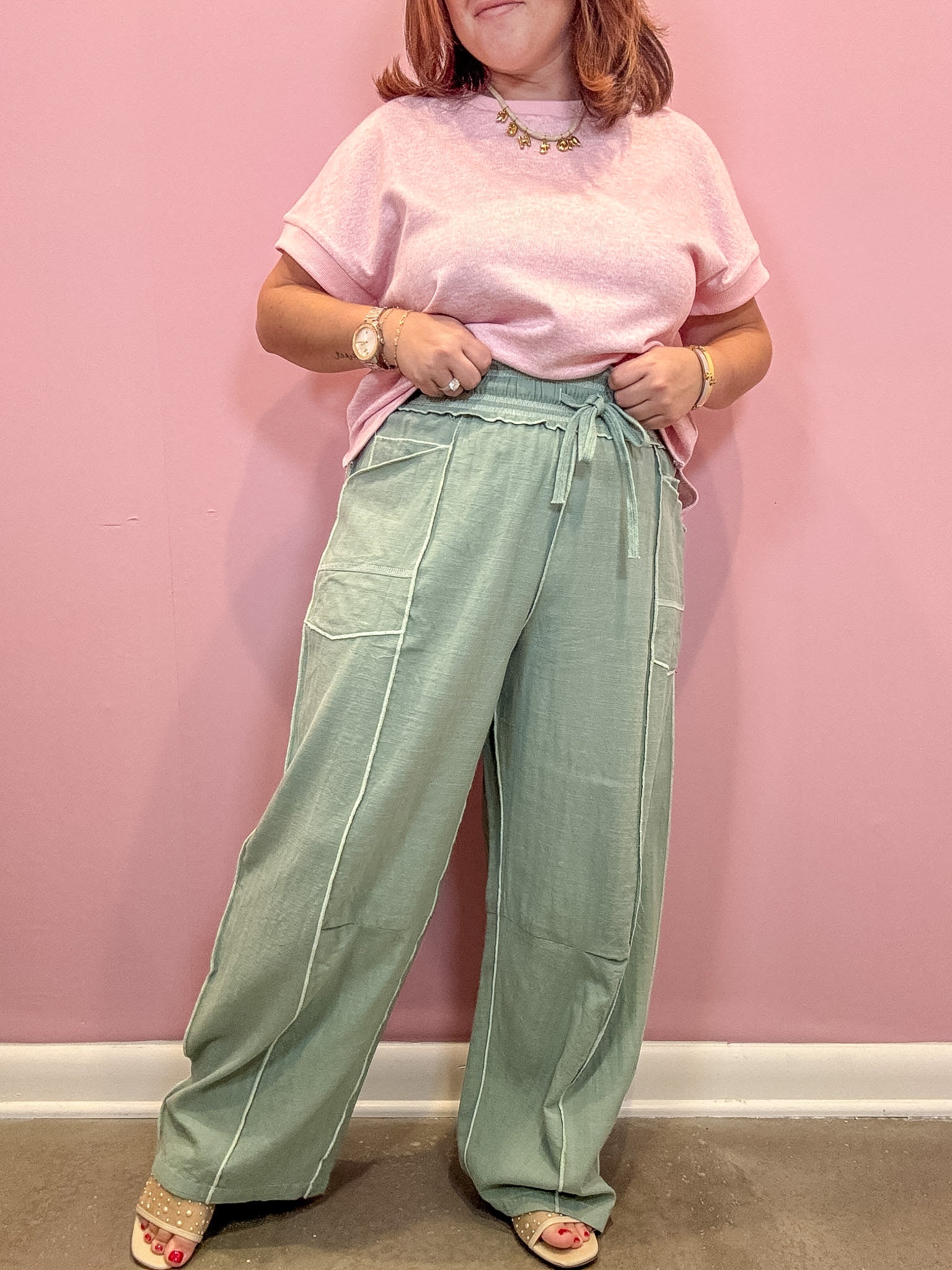 A person wearing a light pink short-sleeve t-shirt and sage green wide-leg drawstring pants, standing against a solid pink wall.