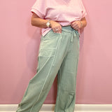 A person wearing a light pink short-sleeve t-shirt and sage green wide-leg pants with white contrast stitching, standing against a solid pink wall.