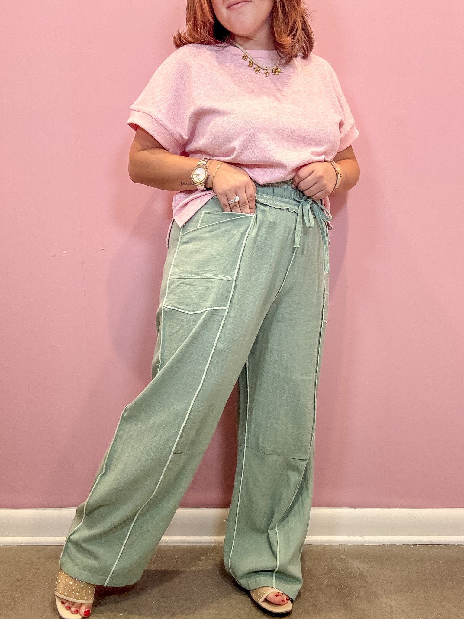 A person wearing a light pink short-sleeve t-shirt and sage green wide-leg pants with white contrast stitching, standing against a solid pink wall.