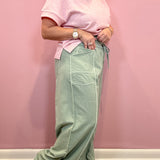 A person wearing a light pink short-sleeve top and sage green wide-leg pants with white contrast stitching, standing against a pink wall.