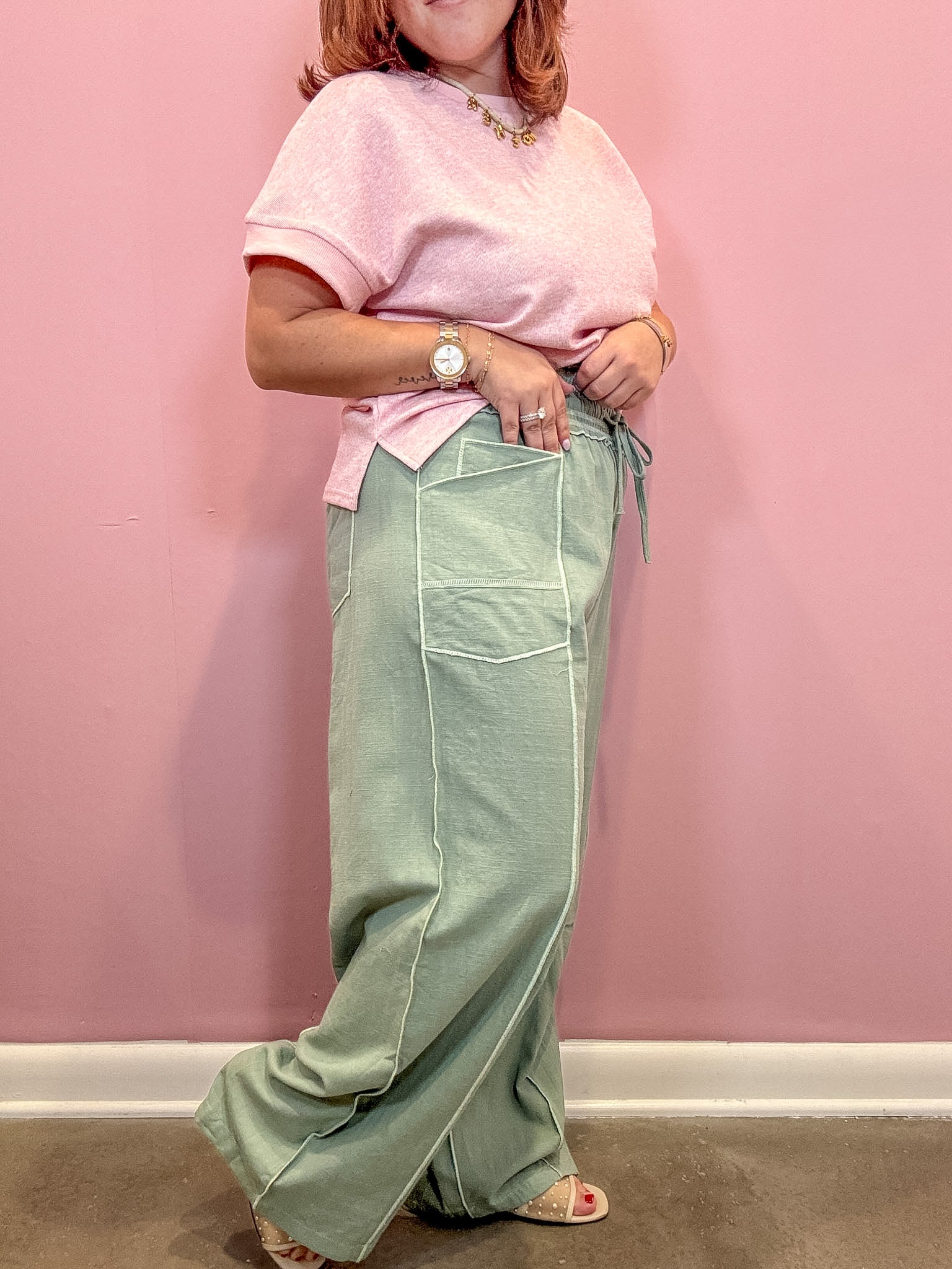 A person wearing a light pink short-sleeve top and sage green wide-leg pants with white contrast stitching, standing against a pink wall.