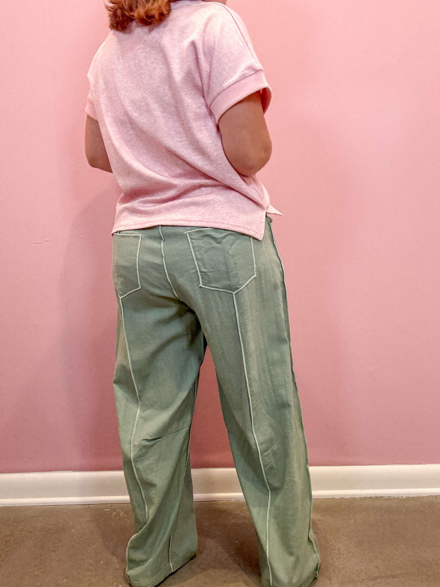 A person shown from the back wearing a light pink short-sleeve t-shirt and sage green wide-leg pants with white contrast stitching, standing against a pink wall.