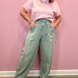 A woman modeling a light pink short-sleeve t-shirt and sage green wide-leg drawstring pants, standing against a solid pink wall.