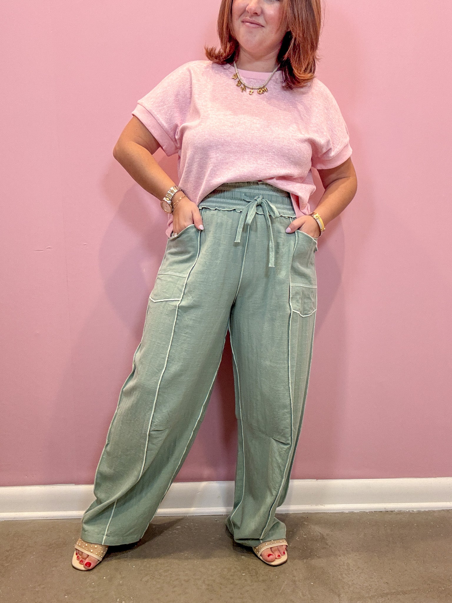 A woman modeling a light pink short-sleeve t-shirt and sage green wide-leg drawstring pants, standing against a solid pink wall.