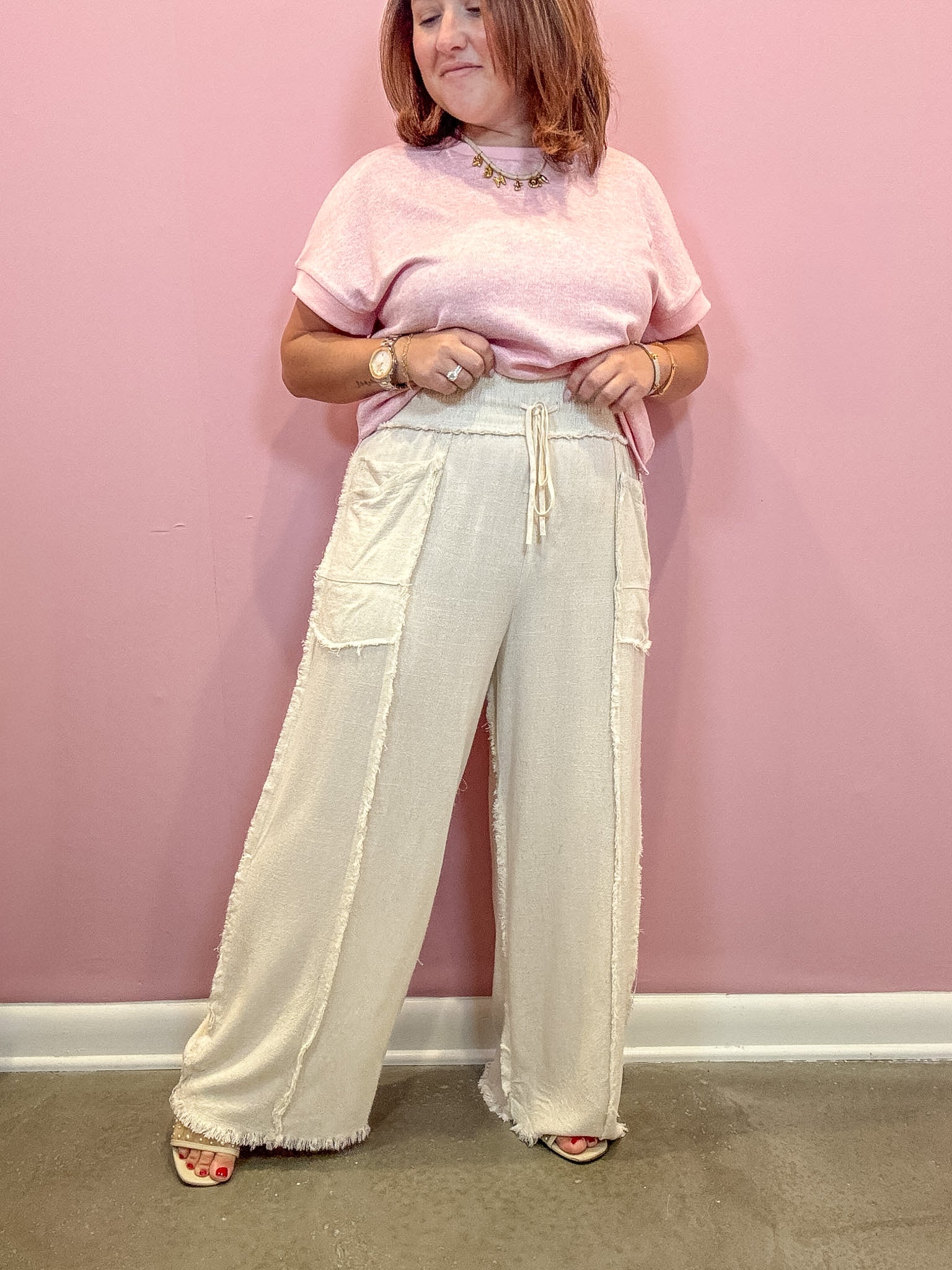 A woman stands against a pink wall wearing a light pink short-sleeve top and cream wide-leg pants with a drawstring waist and frayed hems.