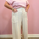 A person wearing a light pink short-sleeve top and off-white wide-leg drawstring pants with large patch pockets, standing against a pink wall.