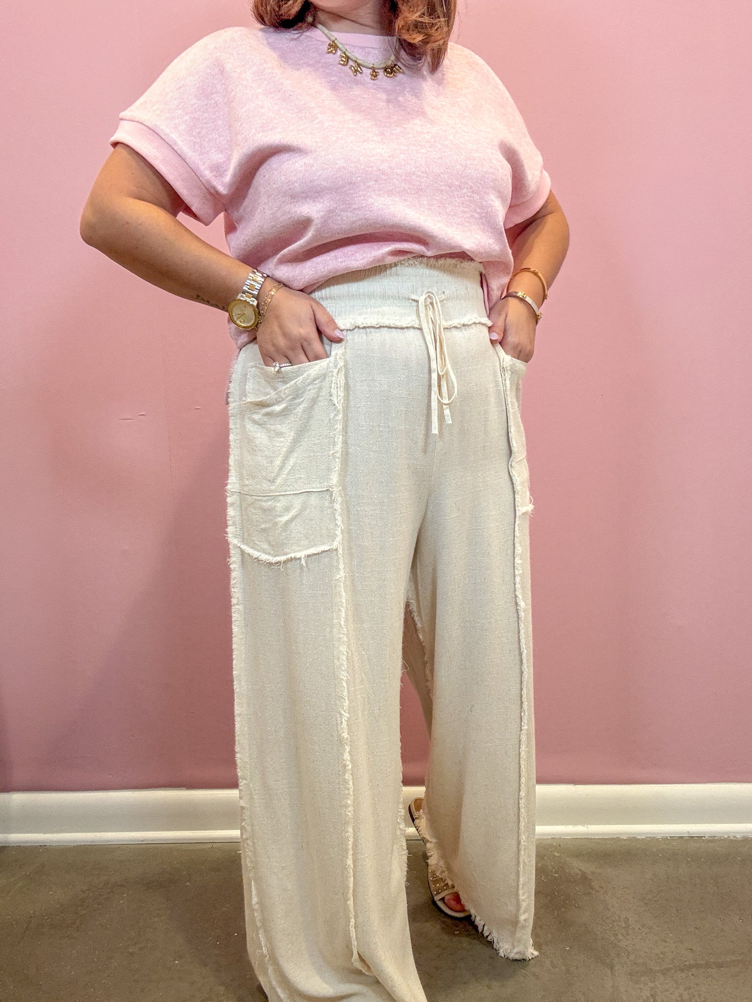 A person wearing a light pink short-sleeve top and off-white wide-leg drawstring pants with large patch pockets, standing against a pink wall.