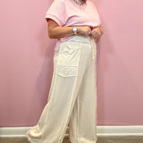 A woman modeling a light pink short-sleeve t-shirt and cream wide-leg linen pants with fringe, standing against a solid pink wall.