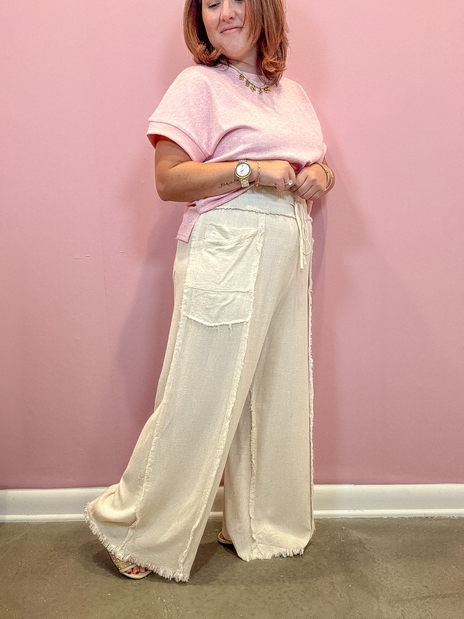 A woman modeling a light pink short-sleeve t-shirt and cream wide-leg linen pants with fringe, standing against a solid pink wall.