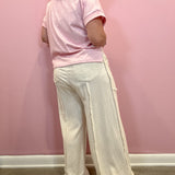 A woman shown from the back wearing a light pink short-sleeve t-shirt and off-white wide-leg pants with raw fringe trim, standing against a solid pink wall.