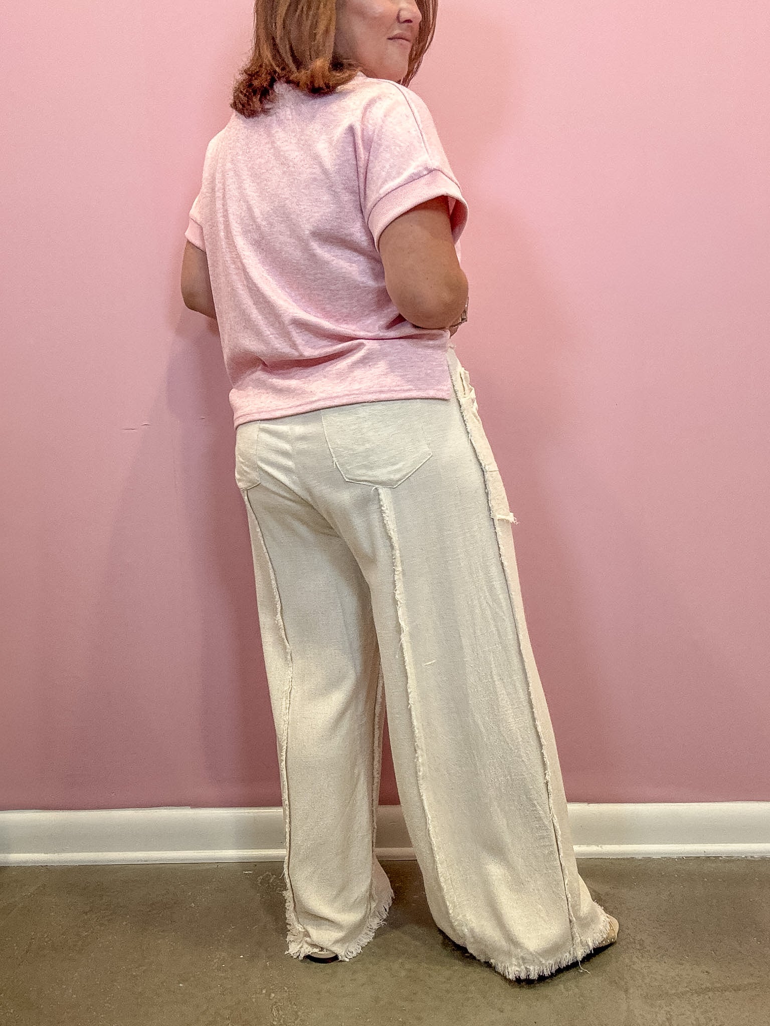 A woman shown from the back wearing a light pink short-sleeve t-shirt and off-white wide-leg pants with raw fringe trim, standing against a solid pink wall.