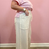 A person wearing a light pink short-sleeve t-shirt and cream wide-leg linen pants with raw edges and a side drawstring, standing against a pink wall.