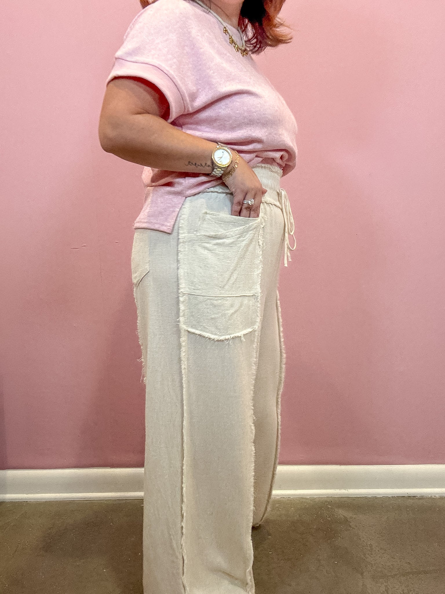 A person wearing a light pink short-sleeve t-shirt and cream wide-leg linen pants with raw edges and a side drawstring, standing against a pink wall.