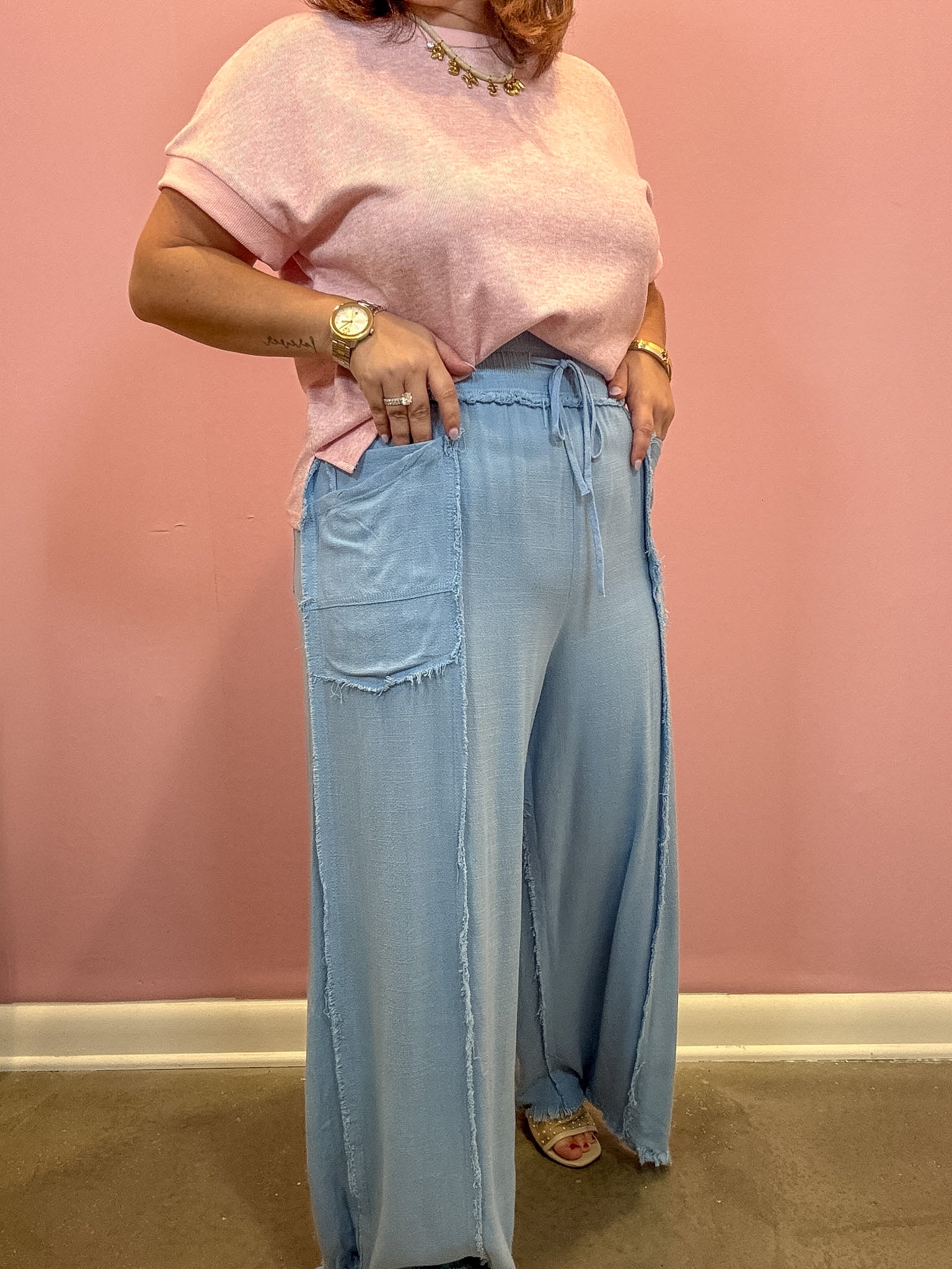 A person wearing a light pink short-sleeve knit top and light blue wide-leg denim pants with raw hems, standing against a pink wall.